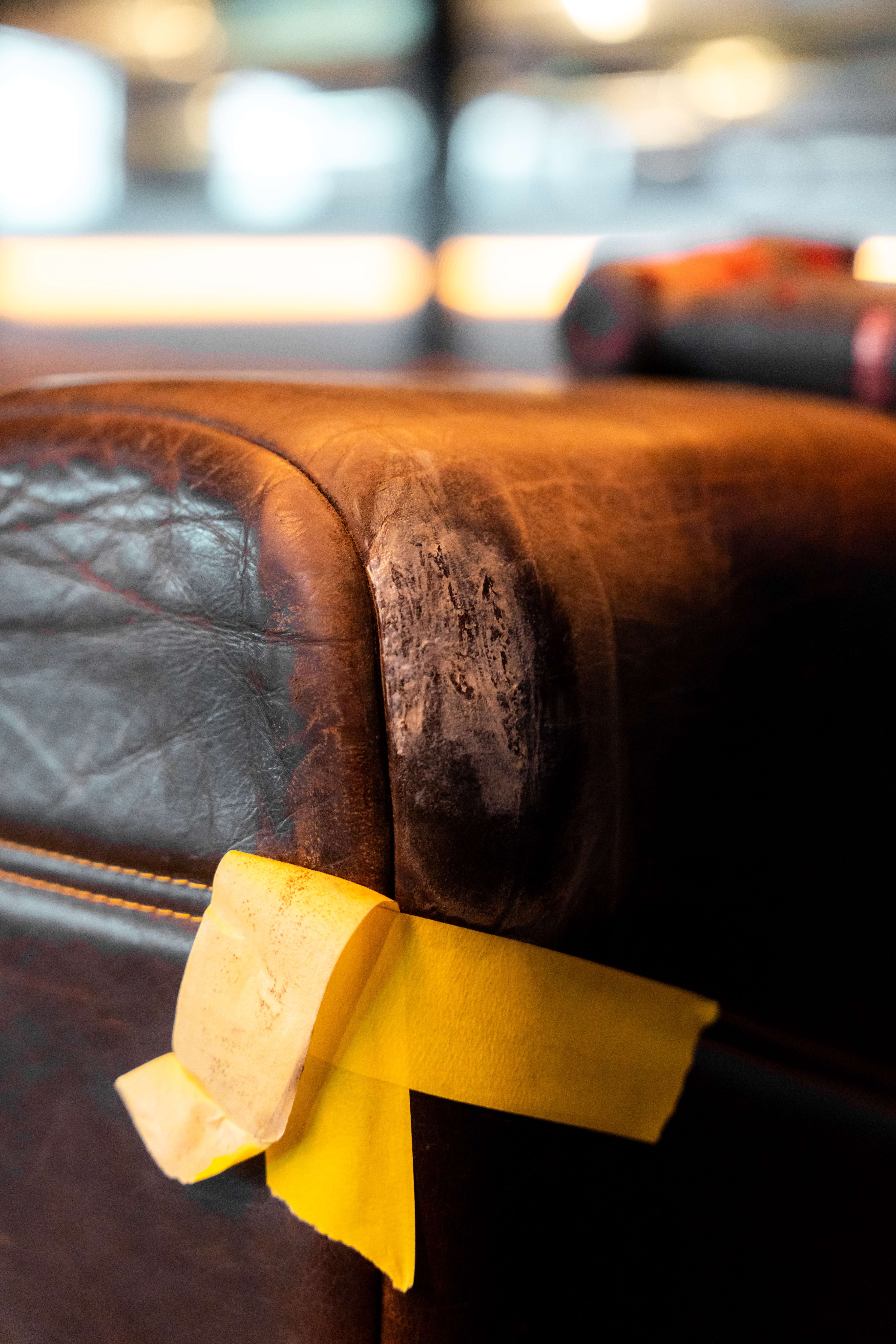 Close up of a dark brown leather lounge arm with a large scuffed patch, masking tape applied along the edge ready for repair