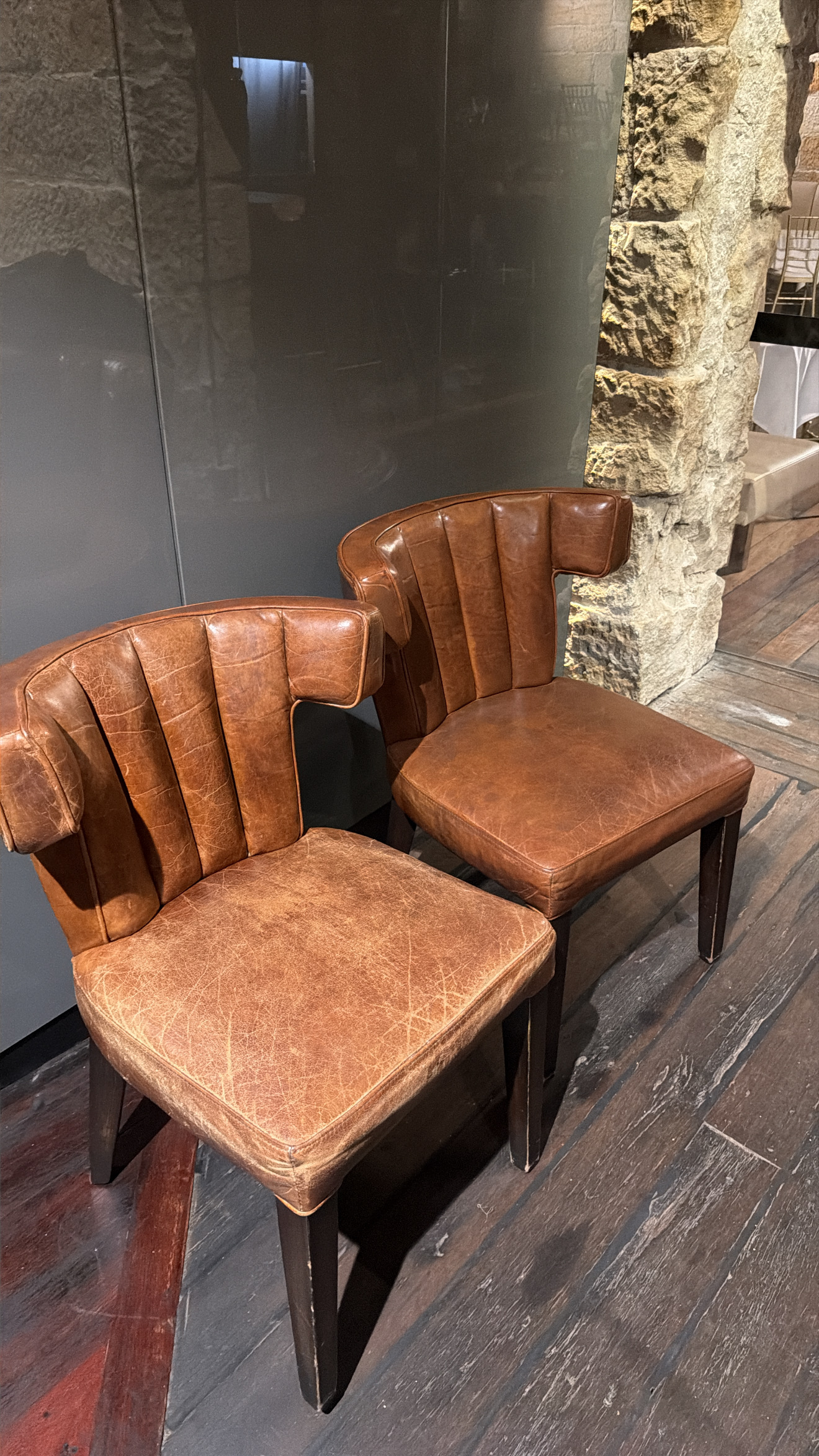 Two matching tan leather channel back dining chairs side by side in a restaurant. The front chair has a scratched, faded seat. The chair behind it has been restored to an even colour