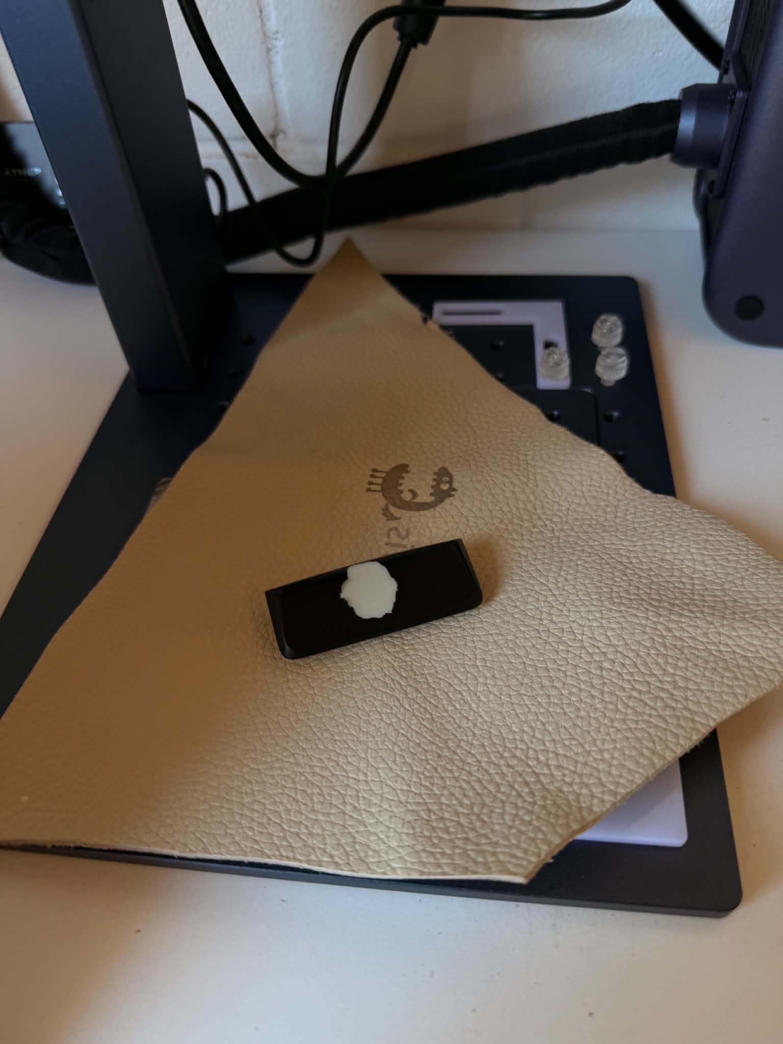 A failed black button on the laser bed with the sticky soft touch coating bubbling and lifting off its face, sitting on a leather test swatch
