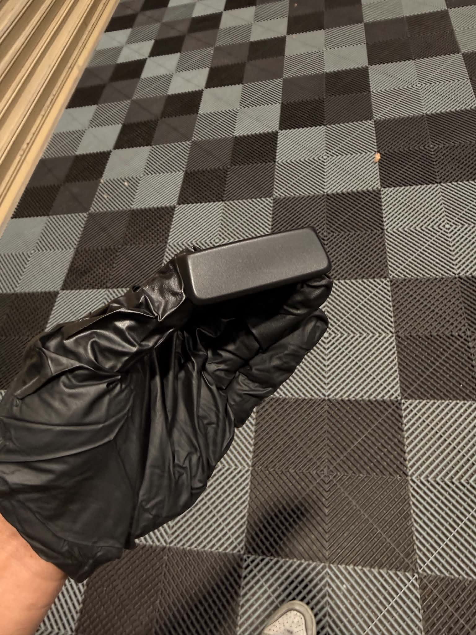 A black plastic trim piece held in a gloved hand on the workshop floor, ready for laser texturing